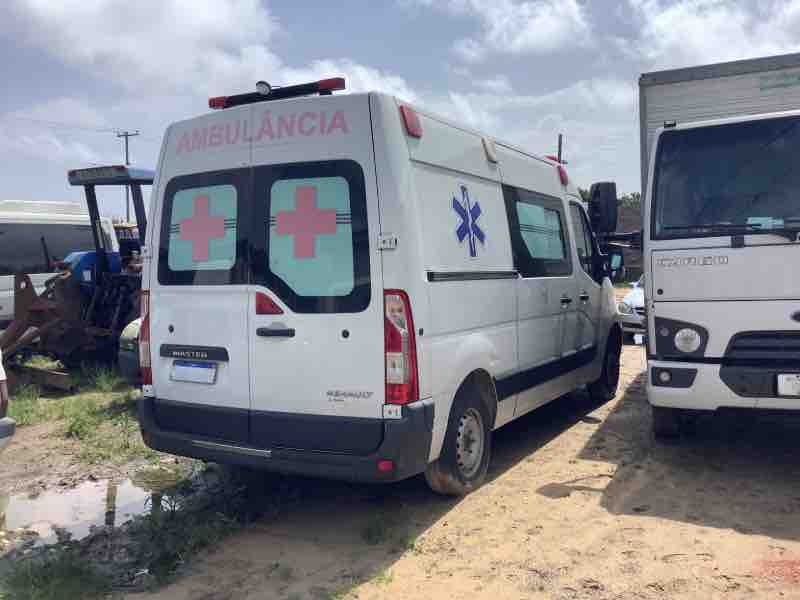 Ambulância branca estacionada em local de armazenamento, com sinais de uso intenso, como parte da inspeção de bens inservíveis para o poder público.
