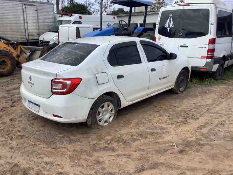 Carro Renault Logan branco, estacionado em área aberta, em estado inservível, com aparência desgastada e suja, como parte do processo de avaliação de bens móveis inservíveis para o poder público.