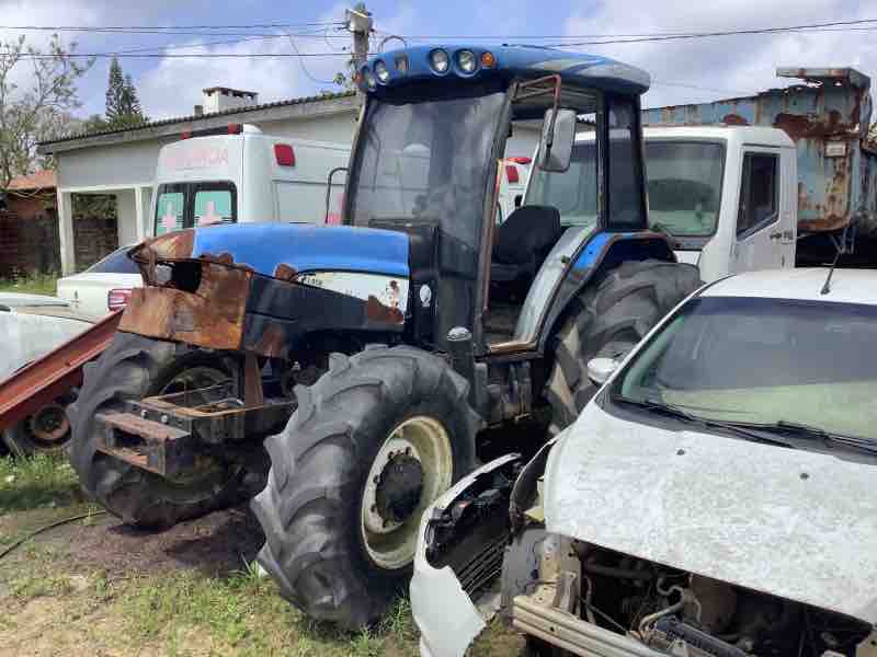 Trator agrícola azul com avarias visíveis, incluindo ferrugem e falta de peças, em local de avaliação de bens móveis inservíveis para o poder público.