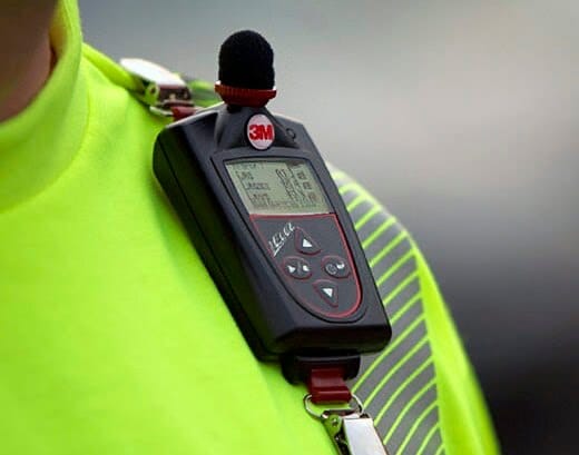 Dosímetro de ruído da 3M preso ao colete de um trabalhador, representando serviços de laudo de ruído ocupacional para análise de exposição sonora e saúde auditiva.