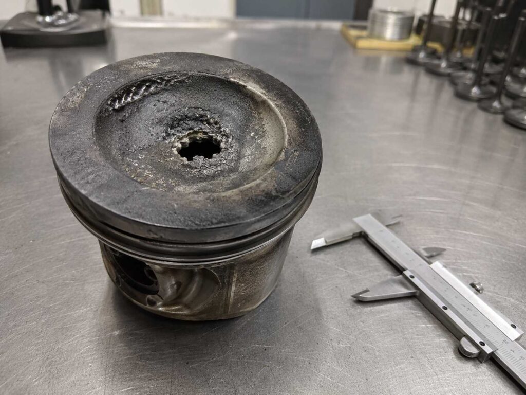 Fotografia forense de um pistão furado em motor diesel com erosão térmica no centro da cabeça do pistão, analisado em perícia de engenharia forense na comarca de Garibaldi, RS.