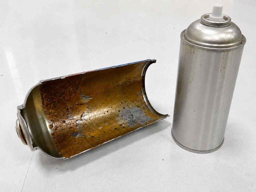 Fotografia técnica de perícia em lata de aerossol em Novo Hamburgo RS, com destaque para a fenda longitudinal e o revestimento de epóxi fenólico com pontos visíveis de corrosão.
